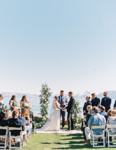 A couple stands at an outdoor wedding ceremony by a lake, surrounded by their bridal party and guests seated on white chairs, with mountains visible in the background.