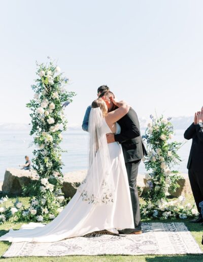 Bride and groom kissing at an outdoor wedding ceremony by the water, surrounded by guests and floral decorations.