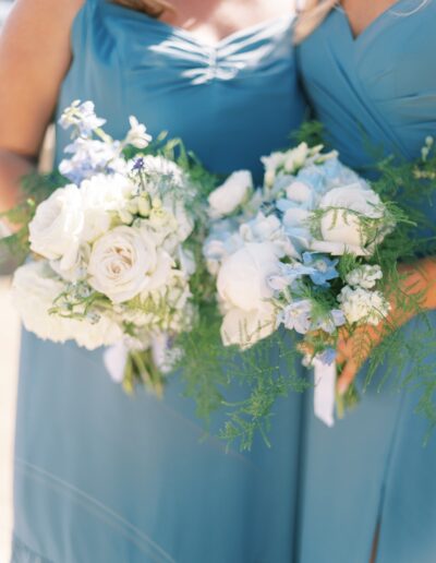 Two people in blue dresses hold bouquets of white and light blue flowers with greenery.