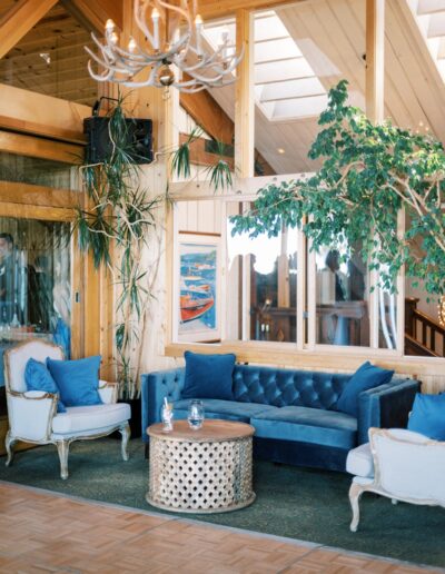 A cozy living room with a blue velvet sofa, two armchairs with blue cushions, a round coffee table, and indoor plants. The room has wooden walls and a chandelier.