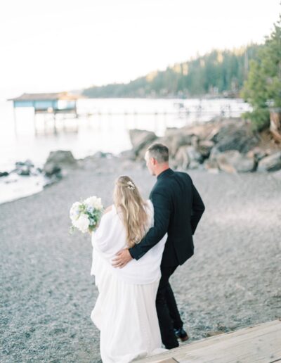 A couple in formal attire walks along a rocky lakeside, with a pier and trees in the background. The woman holds a bouquet.