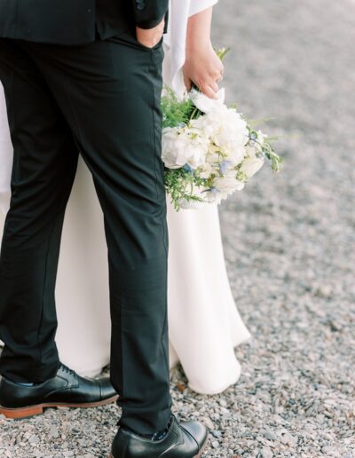 A bride and groom stand on a gravel path. The groom wears a black suit and shoes, while the bride holds a bouquet of white flowers.