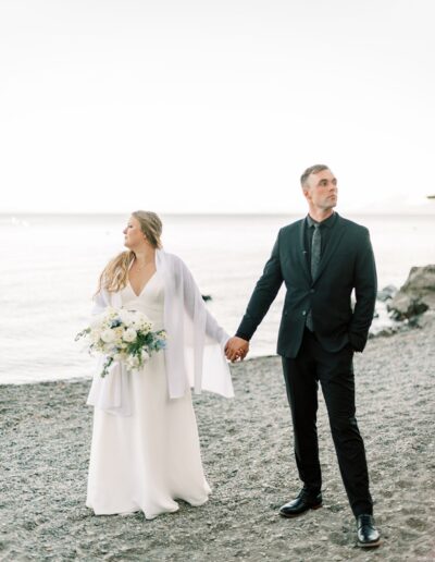 A bride and groom hold hands on a rocky beach, with the bride in a white dress and shawl holding a bouquet, and the groom in a black suit.