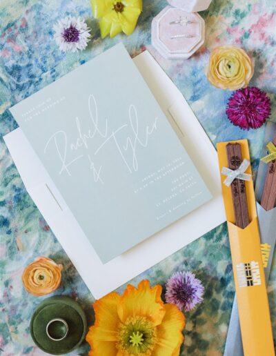 A Napa Valley wedding invitation for "Rachel & Tyler" is surrounded by colorful flowers, a green ribbon, rings in a box, and two bright yellow envelopes with wooden details on a floral background.