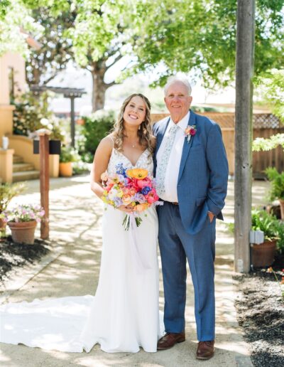 A bride and an older man in formal attire stand outdoors on a sunny Napa Valley day, smiling at the camera. She holds a colorful bouquet amidst the lush greenery.