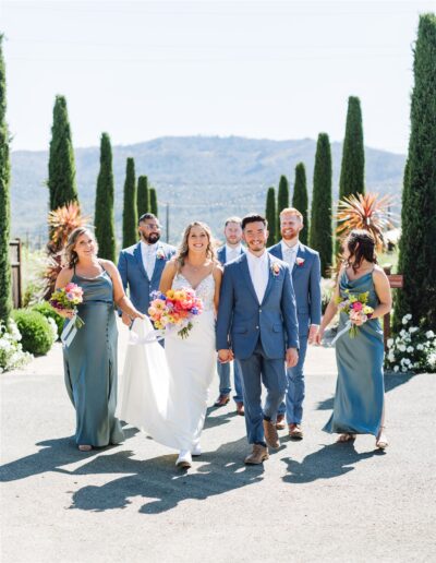 A bride and groom in formal attire walk hand in hand, accompanied by bridal party members holding bouquets. The group is outdoors, surrounded by the lush beauty of a Napa Valley wedding, with tall trees and majestic mountains as their backdrop.