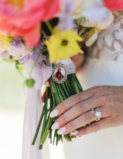 A hand with a ring holds a bouquet of colorful flowers, adorned with a cameo-style brooch featuring a person, capturing the timeless elegance of a Napa Valley wedding.