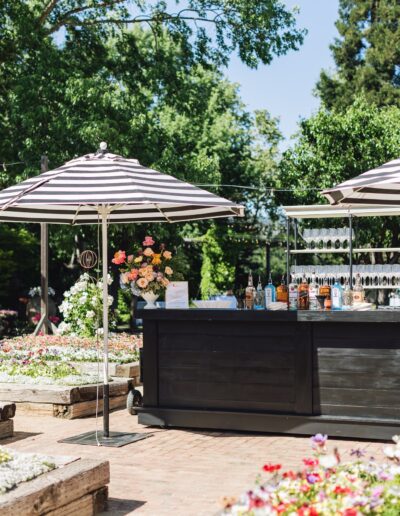 Outdoor bar setup with striped umbrellas, various bottles, and glasses, perfect for a Napa Valley wedding. Surrounded by colorful flower beds and lush greenery, it offers an enchanting setting to celebrate love amidst nature's beauty.