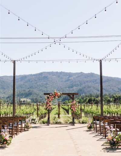 Outdoor Napa Valley wedding setup with wooden chairs and floral arrangements facing an arbor, set against a backdrop of vineyards and mountains under string lights.