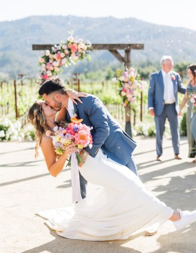 A couple in wedding attire shares a kiss beneath a floral arch, surrounded by onlookers and the breathtaking landscape of a Napa Valley wedding.