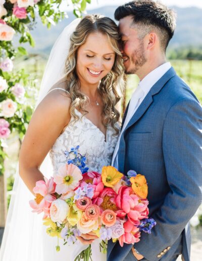 A bride and groom stand closely together outdoors, their love blooming as vibrantly as the colorful bouquet of pink, orange, and yellow flowers she holds. Surrounded by lush greenery and floral decorations, they capture the enchanting romance of a Napa Valley wedding.