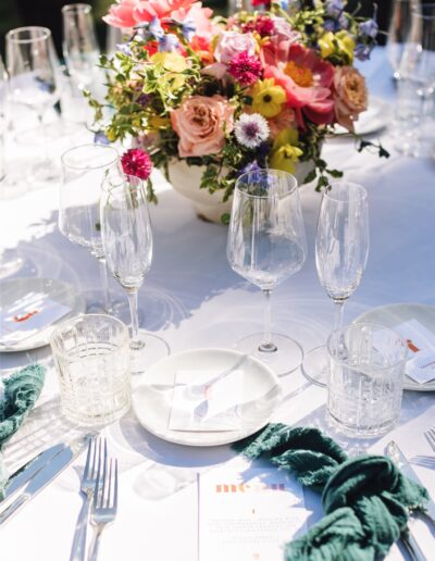 A white tablecloth with dishes, glassware, and green napkins sets the scene for a meal reminiscent of a Napa Valley wedding. A colorful floral arrangement is perfectly centered on the table.