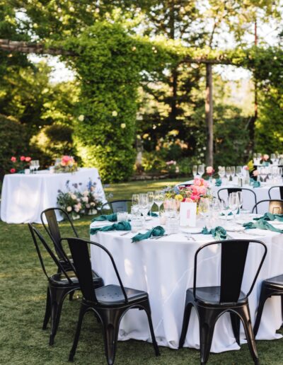 Experience a Napa Valley wedding in an enchanting outdoor garden setting, where round tables draped in white cloth showcase vibrant floral centerpieces and green napkins. Surrounded by black chairs and lush greenery, this picturesque scene captures the essence of a wine country celebration.