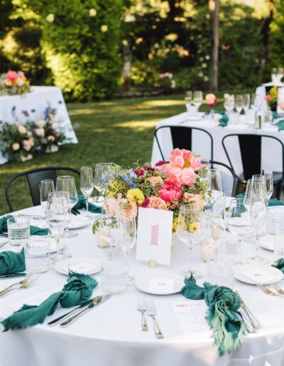 Napa Valley wedding table setting features a white tablecloth, floral centerpiece, glassware, cutlery, and green napkins. A numbered card rests amid the elegance. Lush greenery from the valley is visible in the background, enhancing the serene ambiance.