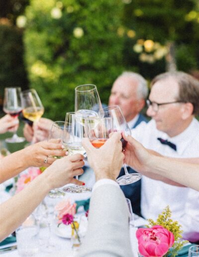 A group of people clinking wine glasses at an outdoor table, surrounded by flowers and greenery, celebrates a picturesque Napa Valley wedding.