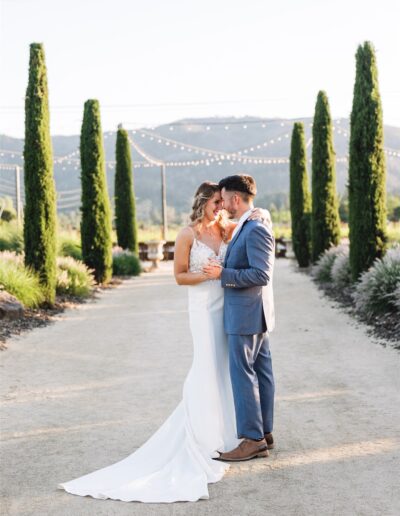 The bride in a white dress and groom in a blue suit stand close on a pathway lined with tall greenery, framed by the string lights and rolling hills of their enchanting Napa Valley wedding.