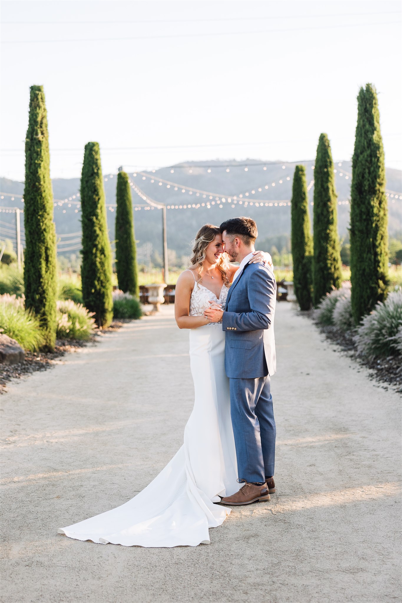 The bride in a white dress and groom in a blue suit stand close on a pathway lined with tall greenery, framed by the string lights and rolling hills of their enchanting Napa Valley wedding.