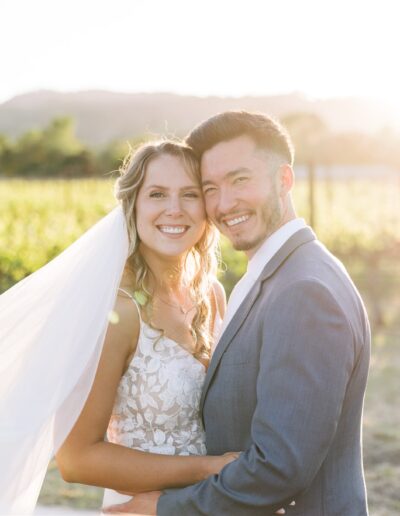 A bride and groom smile while standing in a sunlit Napa Valley vineyard, with the bride's veil flowing in the breeze.