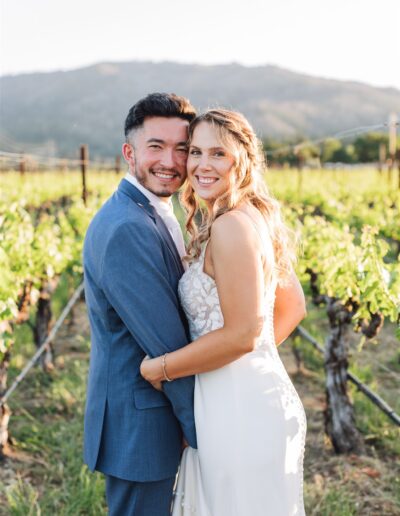 A couple in formal attire embraces in a picturesque Napa Valley vineyard, with rolling hills in the background under a clear sky, capturing the essence of a perfect wedding day.