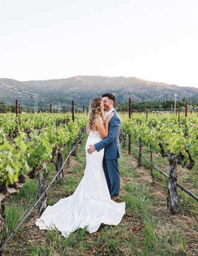 A bride and groom stand embracing in a vineyard, surrounded by rows of grapevines, with the majestic mountains of Napa Valley in the background.