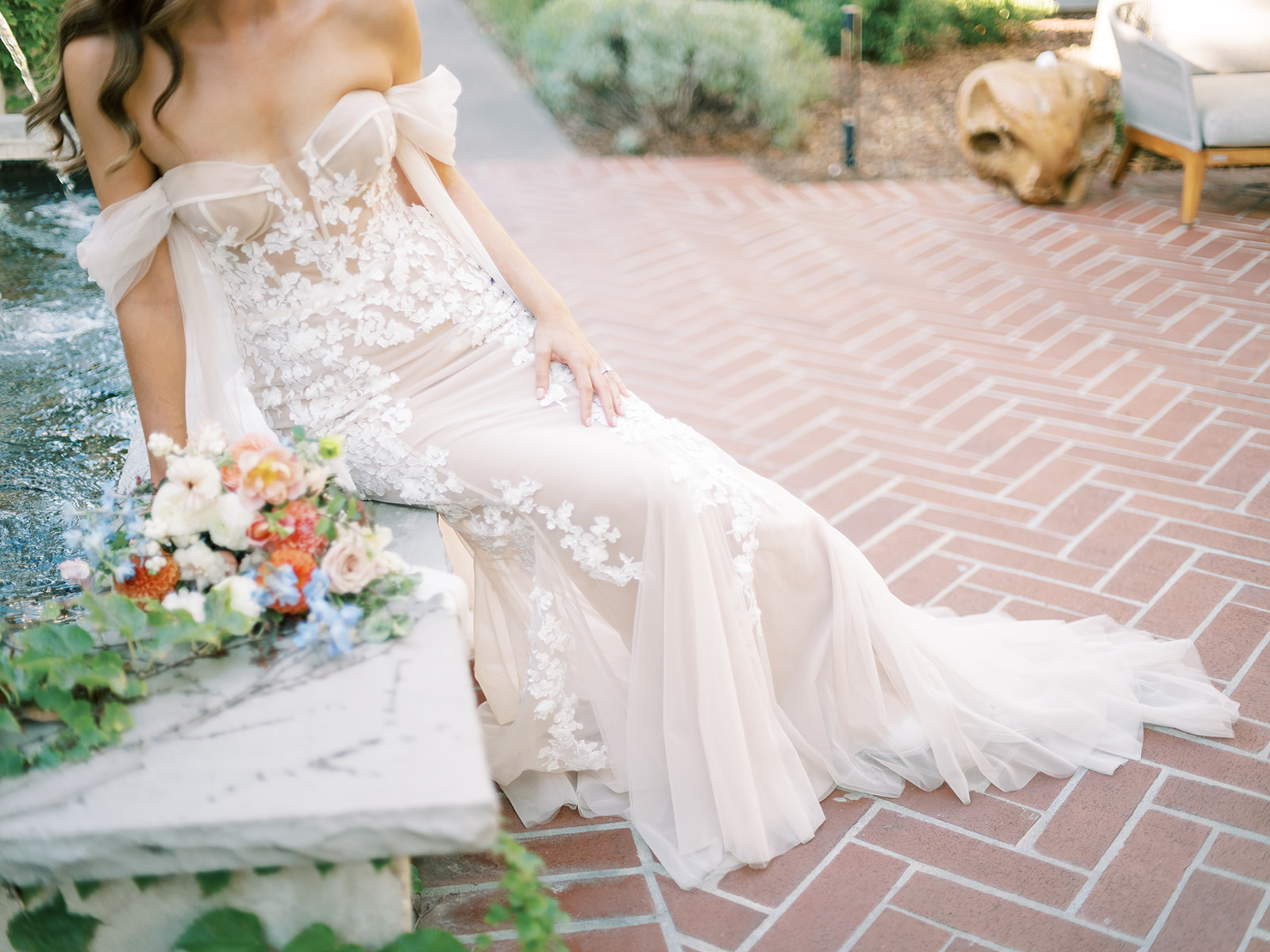 A person in a strapless, lace wedding dress sits on a stone ledge near a fountain, holding a bouquet of colorful flowers. The ground is paved with herringbone brick.