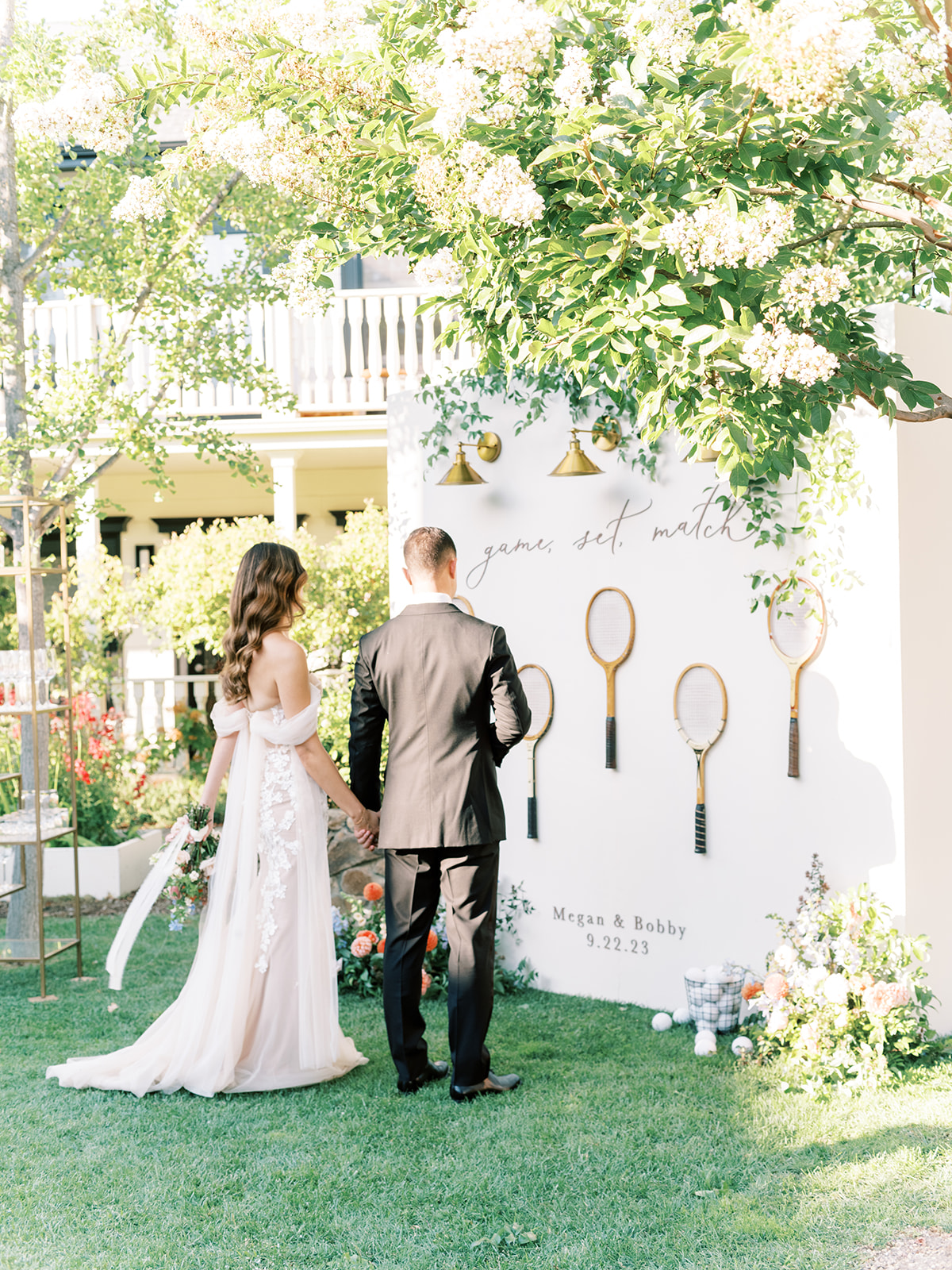 A couple stands holding hands in front of a tennis-themed wedding display with rackets and a sign reading "game, set, match." Lush greenery surrounds the outdoor scene.