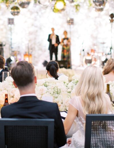 An elegantly dressed crowd seated at tables decorated with white flowers at a wedding reception, listening to speakers on a stage.