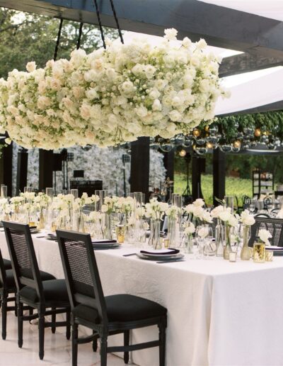 Elegant outdoor wedding reception setup with long tables, white floral centerpieces, black chairs, and large flower arrangement overhead. Tables are covered with white linens.