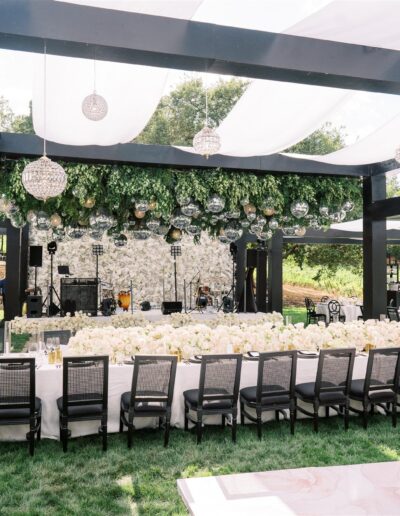 Outdoor reception setup with long dining tables, white tablecloths, and floral arrangements under a black pergola adorned with greenery and hanging orbs. A tiered white cake is in the foreground.