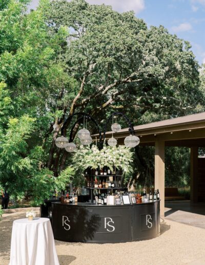 A black outdoor bar stands on a gravel patio under a large tree, with tables nearby. Glasses and drinks are arranged on the bar.