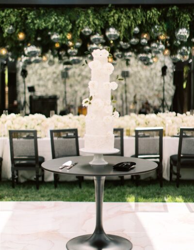 A tall, white, floral-decorated wedding cake on a round black table. Background features a flower-filled banquet setting with hanging greenery and lights.