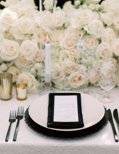 Elegant table setting with white plates, black napkins, and gold accents. Lush white and pale pink roses as a centerpiece.