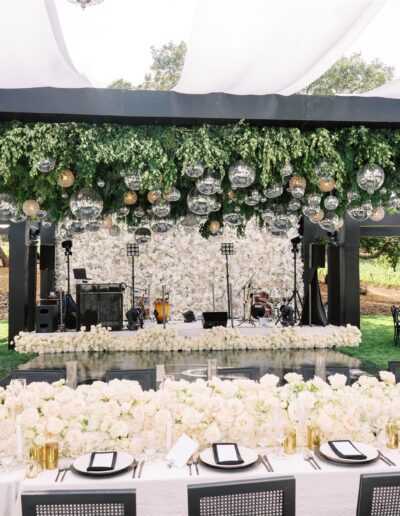 Elegant outdoor wedding setup with a floral-covered stage, hanging glass ornaments, and a long table decorated with white flowers and place settings.