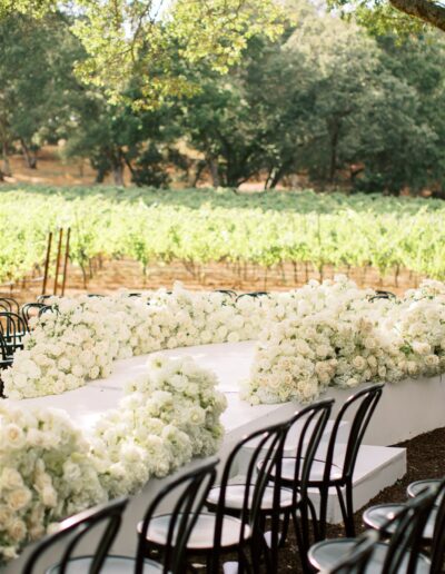 Outdoor wedding setup with chairs arranged around a floral aisle decorated with white roses, set in a vineyard with green trees and grapevines in the background.