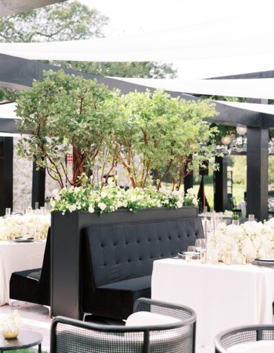 Elegant outdoor event setup with black booths, white floral arrangements, and greenery. Black chairs surround white-clothed tables under a pergola.