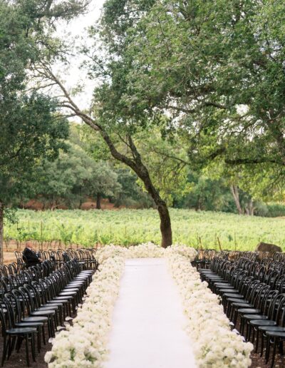 Outdoor wedding aisle lined with white flowers, surrounded by black chairs. Green trees and vineyard visible in the background.