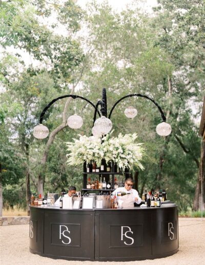 Outdoor bar setup with two bartenders, black counter with "FS" logo, adorned with white flowers and hanging crystal lights, surrounded by trees.