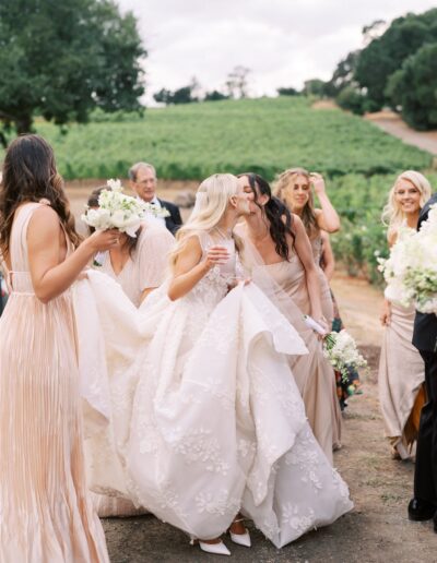 A bride in a white gown shares a kiss with a woman in beige amidst a group of people in formal attire outdoors.