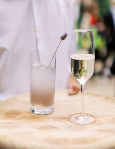 A person holding a wooden tray with a tall glass of iced lavender drink and a champagne flute.