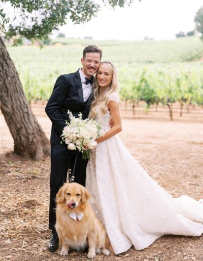 Bride and groom stand smiling with their dog in a vineyard. The bride holds a bouquet, and the dog wears a bow tie.