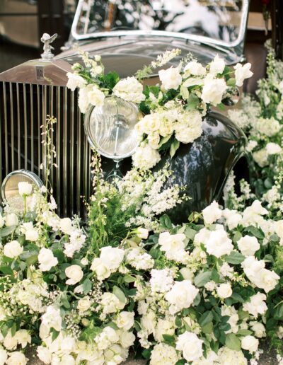 A vintage car adorned with an abundance of white roses and greenery, surrounded by floral arrangements.