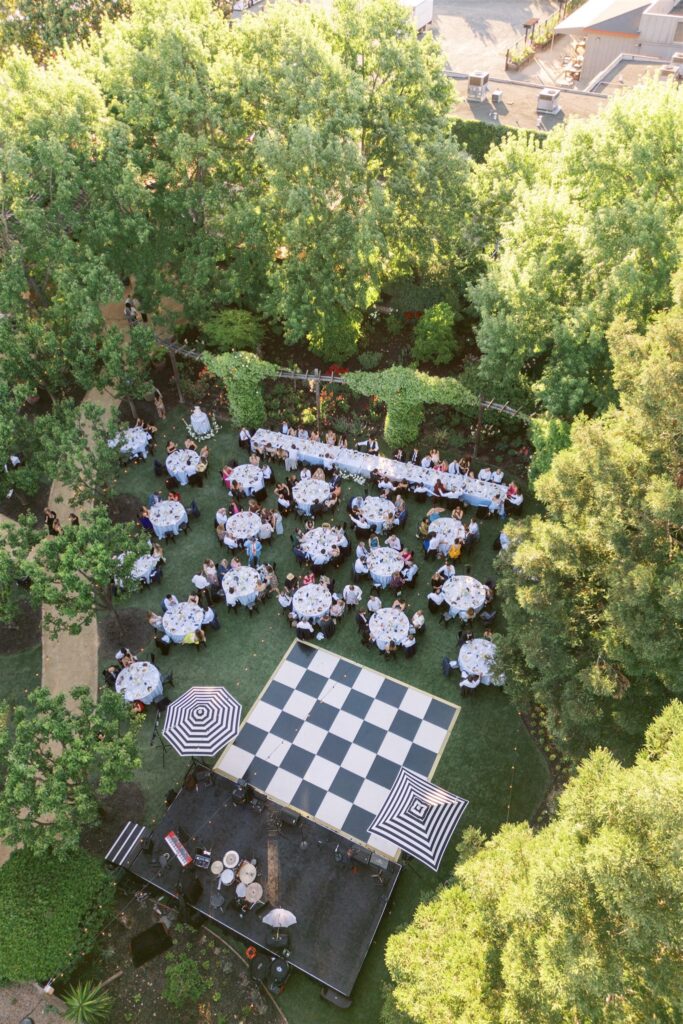 Aerial view of an outdoor event featuring tables set for dining, a large checkered dance floor, and surrounded by green trees.