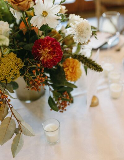 Floral arrangement with red, yellow, and white flowers in a vase on a table, next to lit tea candles.