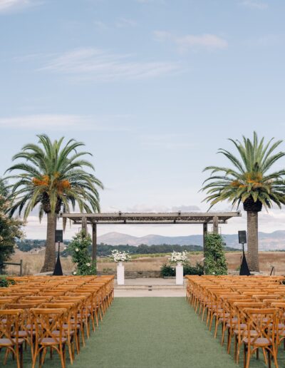 Outdoor wedding setup with rows of wooden chairs facing an altar between two palm trees, with mountains and a blue sky in the background.