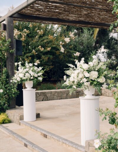 Outdoor wedding ceremony area with two large white floral arrangements on tall pedestals. Stone flooring and greenery in the background.