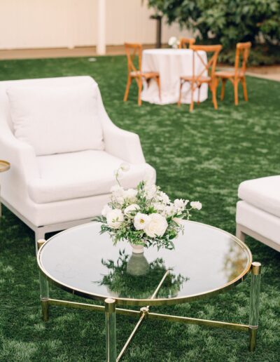 A white armchair is set beside a glass coffee table with a floral arrangement on it, placed on a grassy area. In the background, a round table with a white cloth and chairs is seen.