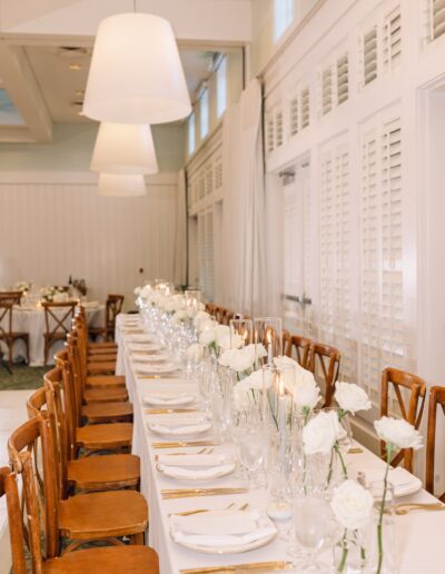 Elegant banquet table setup with white tablecloth, wooden chairs, white roses, glassware, and gold cutlery, in a well-lit room with large pendant lights and white shutters.