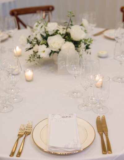 A neatly set table with a white tablecloth features a floral centerpiece, candles, empty wine glasses, and gold utensils. A menu card is placed on a plate.