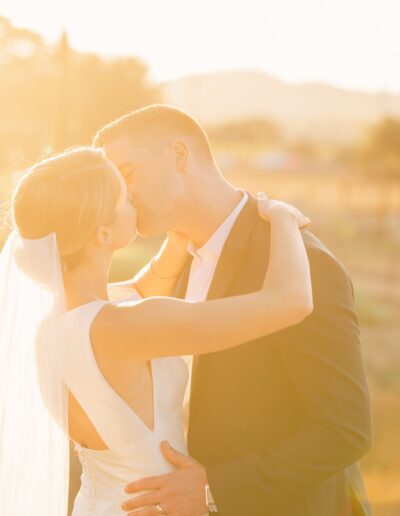 A couple in wedding attire kisses outdoors at sunset, surrounded by warm, golden light.