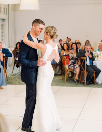 A couple dances at a wedding reception, surrounded by seated guests watching and clapping. The woman wears a white dress; the man is in a dark suit.